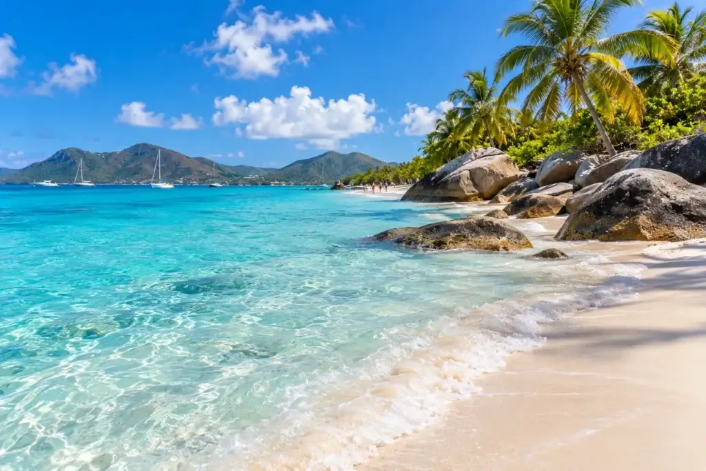 Powder-soft Caribbean beach with clear turquoise water, tropical palms, rocky shoreline, sailboats, and green islands in the distance